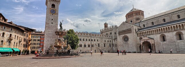 Piazza Duomo a Trento. Sullo sfondo, la Torre Civica, Palazzo Pretorio e la cattedrale di San Vigilio, Duomo della città. Al centro, la fontana del Nettuno. Dietro alla fontana svetta l’unico albero della piazza: un tiglio. La piazza, nell’immagine, è vissuta. Ci sono alcune persone sedute sui gradini della fontana, qualcuno sta scattando delle foto e un gruppetto è in posa davanti alla cattedrale per farsi fotografare. Tutti indossano abiti estivi. Il cielo azzurro è striato dal bianco di nuvole leggere.