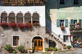 Scorcio di San Lorenzo in Banale in una giornata soleggiata. Nell’immagine spiccano le macchie rosse e viola dei moltissimi vasi di fiori, forse gerani: sono appesi alle finestre e al bordo di una terrazza circondata da archi, appoggiati sui grandini di una scalinata e su un piccolo balcone. Il sole e questi colori vivaci trasmettono un senso di allegria.