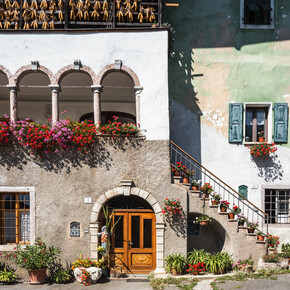 Scorcio di San Lorenzo in Banale in una giornata soleggiata. Nell’immagine spiccano le macchie rosse e viola dei moltissimi vasi di fiori, forse gerani: sono appesi alle finestre e al bordo di una terrazza circondata da archi, appoggiati sui grandini di una scalinata e su un piccolo balcone. Il sole e questi colori vivaci trasmettono un senso di allegria.